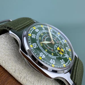 Puede incluir: Un reloj de pulsera de estilo militar con esfera verde y correa de lona verde. El reloj tiene una caja plateada y presenta un s&iacute;mbolo de radiaci&oacute;n. La esfera muestra la hora con n&uacute;meros blancos y amarillos.