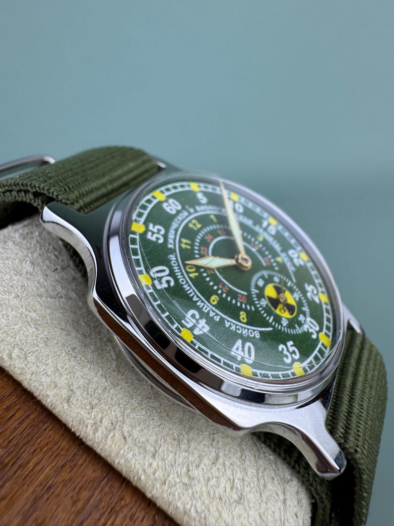 Puede incluir: Un reloj de pulsera de estilo militar con esfera verde y correa de lona verde. El reloj tiene una caja plateada y presenta un s&iacute;mbolo de radiaci&oacute;n. La esfera muestra la hora con n&uacute;meros blancos y amarillos.