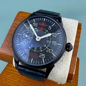 May include: Black wristwatch with a dark blue face, featuring white numerals and hands. The watch has a black leather band with red stitching. The face includes the text "ВОЕННО ВОЗДУШНАЯ РАЗВЕДКА" and a red star and airplane graphic.