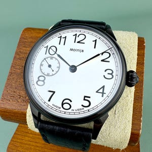 May include: A black Molnija wristwatch with a white face and black numerals. The watch has a small seconds dial and a black leather strap. The words "Molnija" and "Made in USSR" are visible on the watch face.