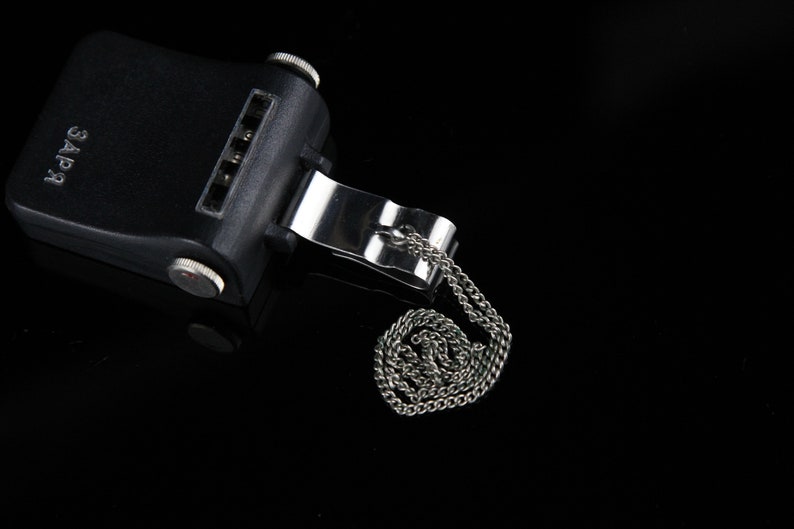 Pode incluir: Um dispositivo preto com um clipe e uma corrente prateados. O dispositivo tem um bot&atilde;o vermelho e a palavra "ЗАРЯ" impressa nele.