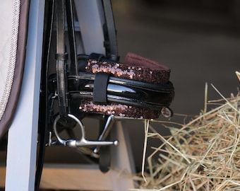 May include: A black leather horse bridle with a brown sequined browband. The bridle is hanging on a white wall.