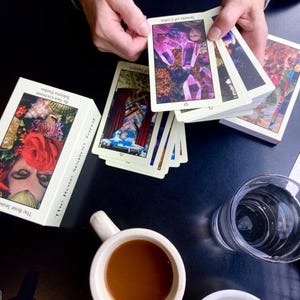 May include: A deck of tarot cards with the text "Seven of Coins" and "The Rose Season Tarot" on the box. The cards are being held by a person. A cup of coffee, a glass of water, and a small bowl with butter are also on the table.