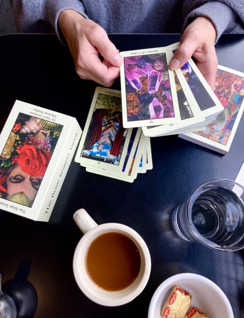 May include: A deck of tarot cards with the text "Seven of Coins" and "The Rose Season Tarot" on the box. The cards are being held by a person. A cup of coffee, a glass of water, and a small bowl with butter are also on the table.