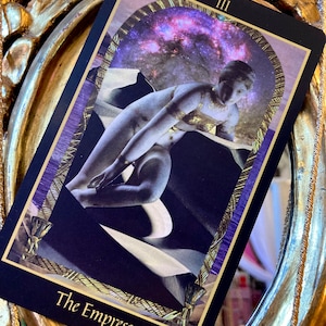 Pu&ograve; includere: La carta dei tarocchi L'Imperatrice con un bordo viola e dorato. La carta presenta una donna con una corona di stelle, seduta su un trono con una luna crescente dietro di lei. Il numero della carta III &egrave; nell'angolo in alto a destra.