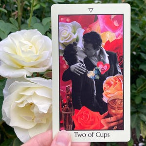 May include: A tarot card with the text "Two of Cups" features a collage of a kissing couple, roses, and a cocktail. The card has a white border and a triangle at the top. The background includes white and red roses.