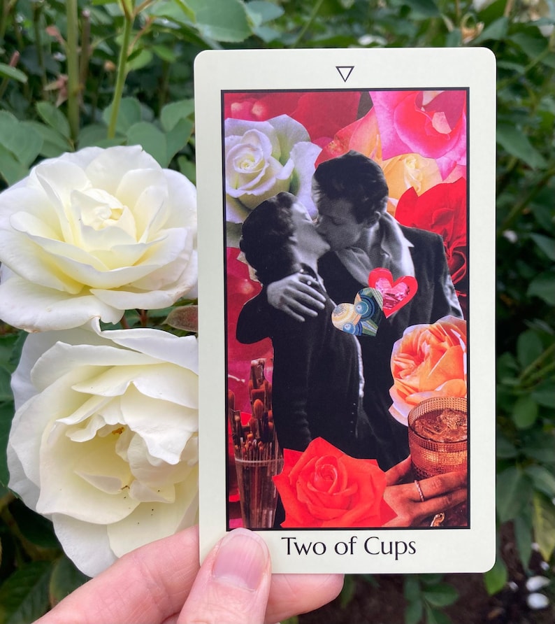 May include: A tarot card with the text "Two of Cups" features a collage of a kissing couple, roses, and a cocktail. The card has a white border and a triangle at the top. The background includes white and red roses.