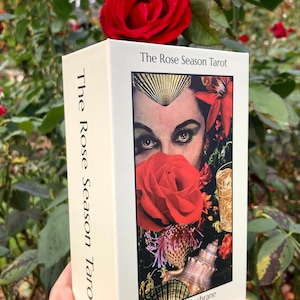 May include: A tarot card deck titled "The Rose Season Tarot" with a cream-colored box. The box features an image of a woman's face, a red rose, and other floral elements. The deck is held in front of a background of green foliage and red roses.