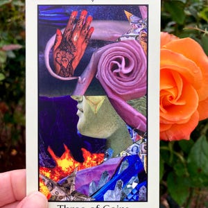 May include: A tarot card with a collage design featuring a hand with henna, a pink spiral, and a profile with a gold triangle. The card reads "Three of Coins" at the bottom. An orange rose is in the background.