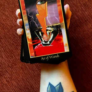A hand holds a deck of Tarot cards showing the Ace of Wands on top. The card features a stylized bolt of orange lightning zapping between a human eye and a feline mouth. In the background, a lightning storm rages above a city.