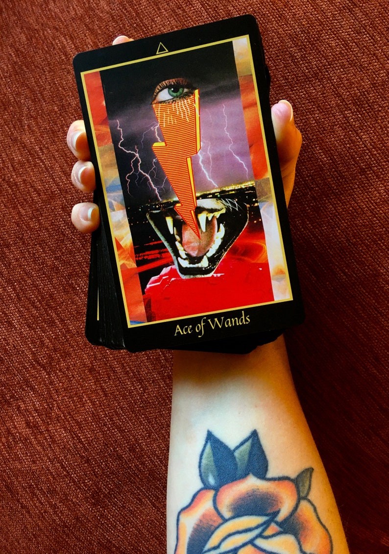 A hand holds a deck of Tarot cards showing the Ace of Wands on top. The card features a stylized bolt of orange lightning zapping between a human eye and a feline mouth. In the background, a lightning storm rages above a city.