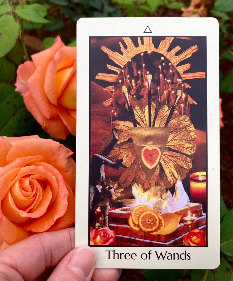 May include: A tarot card titled "Three of Wands" featuring a collage with a golden sunburst, a heart-shaped fruit, and orange slices. The card is held near two orange roses.