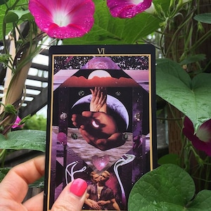 The Lovers card from the Telezma Tarot is seen held by a hand in front of blooming pink morning glories. On the card, two male lovers embrace beneath a celestial geometric composition including the Earth as seen from the Moon.