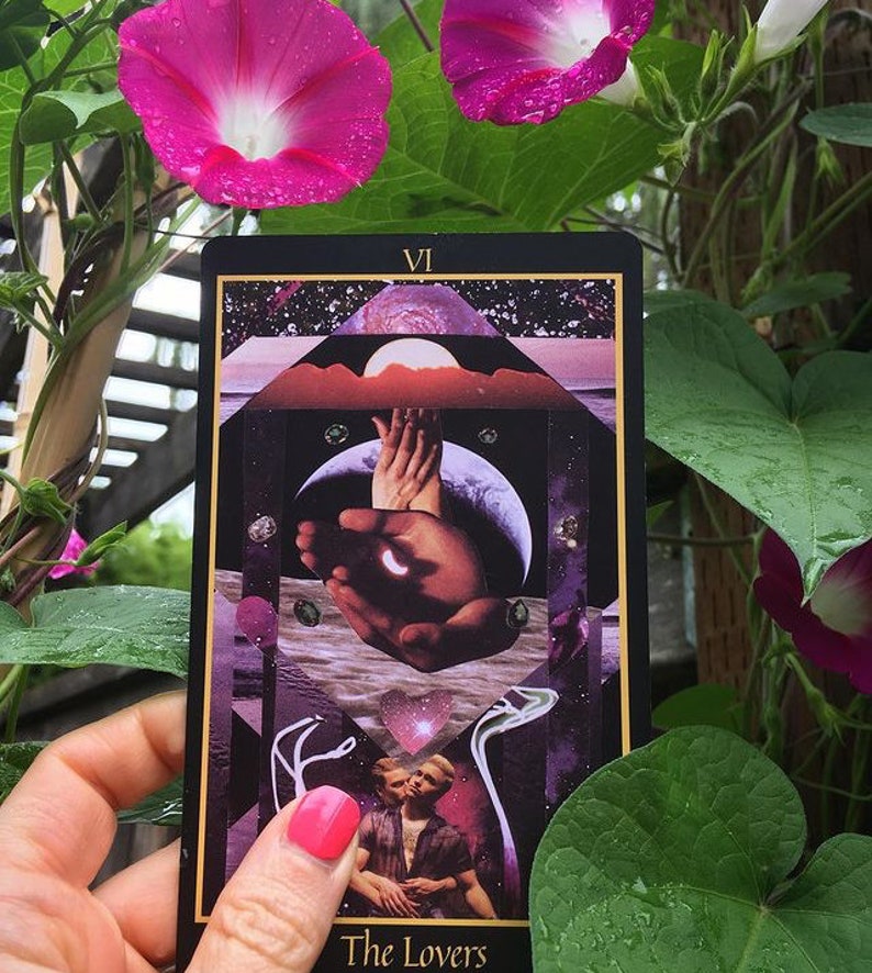 The Lovers card from the Telezma Tarot is seen held by a hand in front of blooming pink morning glories. On the card, two male lovers embrace beneath a celestial geometric composition including the Earth as seen from the Moon.