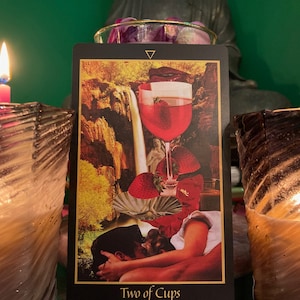 Pu&ograve; includere: Lettura di carte dei tarocchi con una candela accesa e una carta Due di Coppe. La carta mostra una coppia che si abbraccia con una cascata e un bicchiere di vino sullo sfondo. Il testo della carta recita "Due di Coppe".