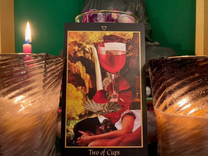 Pu&ograve; includere: Lettura di carte dei tarocchi con una candela accesa e una carta Due di Coppe. La carta mostra una coppia che si abbraccia con una cascata e un bicchiere di vino sullo sfondo. Il testo della carta recita "Due di Coppe".