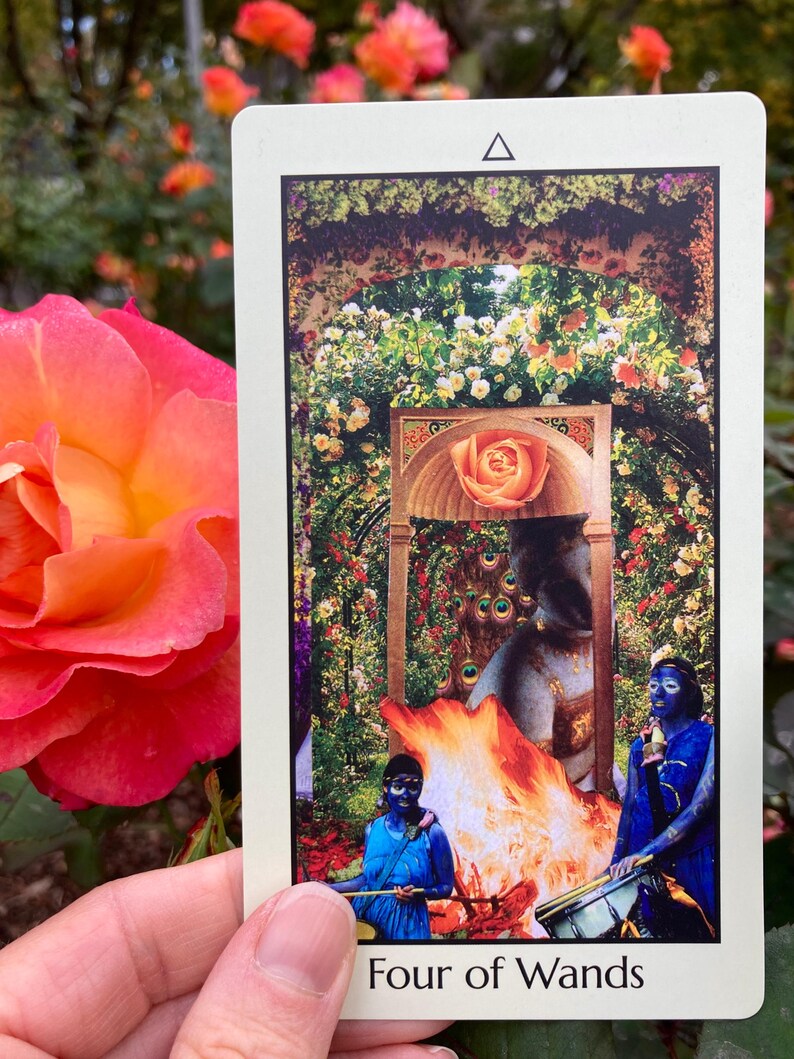 May include: A tarot card with a floral and mythological design. The card features a central image of a rose, figures in blue, and a fire. The text "Four of Wands" is at the bottom. The card is held in front of a background of roses.