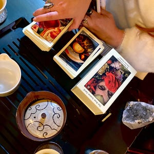 May include: A tarot card reading setup featuring a deck of The Rose Season Tarot cards, with the word "Fortune" visible on one card. The scene includes a tea set with a decorative ceramic pot and small cups, and a crystal.