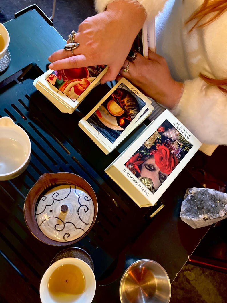 May include: A tarot card reading setup featuring a deck of The Rose Season Tarot cards, with the word "Fortune" visible on one card. The scene includes a tea set with a decorative ceramic pot and small cups, and a crystal.
