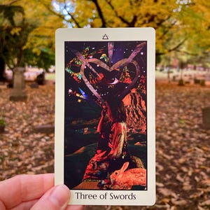 May include: A tarot card titled "Three of Swords" is held in front of a blurred outdoor background. The card features a collage-style illustration with a heart shape, a figure, and a rabbit. The top of the card has a triangle symbol.