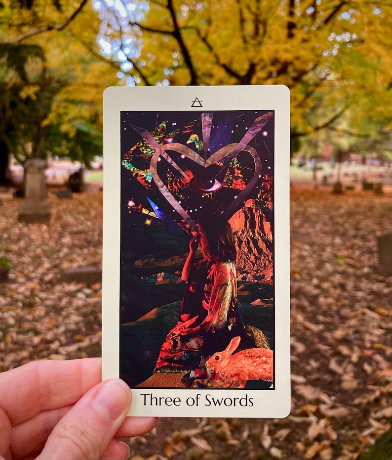 May include: A tarot card titled "Three of Swords" is held in front of a blurred outdoor background. The card features a collage-style illustration with a heart shape, a figure, and a rabbit. The top of the card has a triangle symbol.