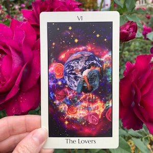 May include: A tarot card featuring a cosmic scene with Earth, roses, and two figures embracing. The card is labeled "VI" at the top and "The Lovers" at the bottom. The background includes a vibrant nebula and scattered stars. The card is held in front of a backdrop of red roses.