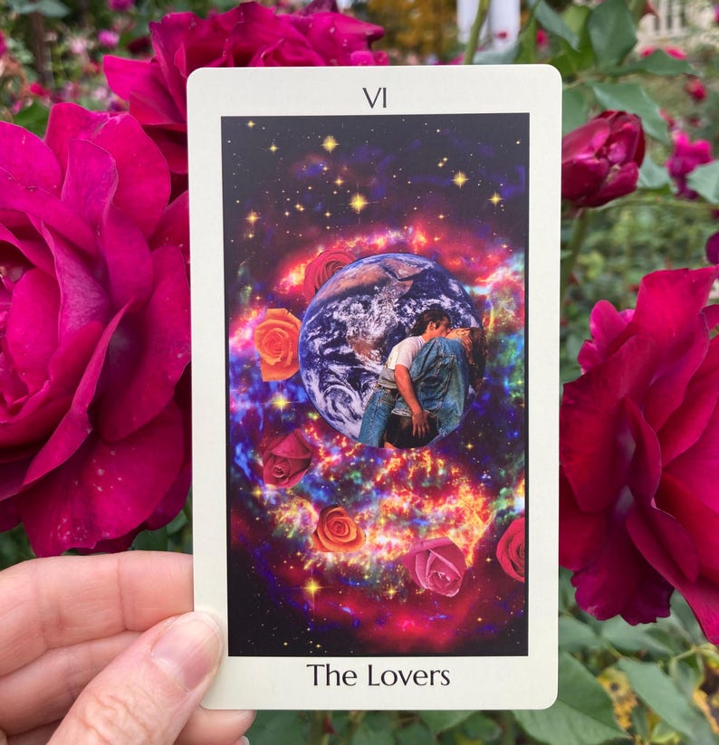 May include: A tarot card featuring a cosmic scene with Earth, roses, and two figures embracing. The card is labeled "VI" at the top and "The Lovers" at the bottom. The background includes a vibrant nebula and scattered stars. The card is held in front of a backdrop of red roses.