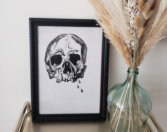 Puede incluir: Dibujo en blanco y negro de un cráneo con lágrimas que gotean, enmarcado en un marco negro. La obra de arte se exhibe en una mesa con un jarrón de hierba de la pampa seca.