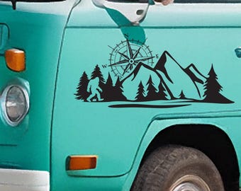Könnte beinhalten: Schwarzer Vinyl-Aufkleber mit einer Bergkette, einer Windrose, einem Bigfoot und Kiefern. Der Aufkleber ist an der Seite eines türkis farbenen Fahrzeugs angebracht, in der Nähe des Vorderrads und des Scheinwerfers.
