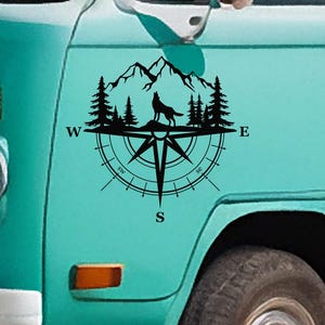 Puede incluir: Un diseño de brújula en blanco y negro con un lobo aullando a las montañas. La brújula está rodeada de árboles y las palabras "W", "E", "S", "SW" y "SE". El diseño está en el lateral de una furgoneta azul turquesa.