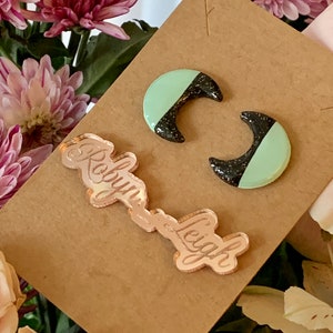 May include: A pair of crescent moon shaped earrings with black and mint green glitter. The earrings are displayed on a brown card with the name "Roun-Leigh" written in gold.