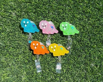 May include: Five colorful dinosaur-shaped badge reels. The dinosaurs are in shades of blue, pink, green, orange, and yellow. Each dinosaur has a glittery finish and is attached to a retractable reel with a clip.