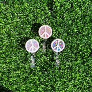 May include: Three peace sign shaped badge reels with glitter accents. The badge reels are pink, white, and multi-coloured.