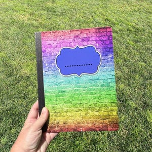 May include: A rectangular notebook featuring a vibrant rainbow-colored brick wall pattern across the cover. A solid purple, scalloped-edge label with a dotted line for writing is centered on the front. The notebook has a black spine binding.