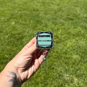 Op de afbeelding: Een vierkante badgehouder met een glinsterende lichtgroene achtergrond en zwarte tekst die CAUTION: OVERSTIMULATED DO NOT APPROACH leest. De badge is omlijst door overlappende zwarte vierkante contouren en heeft een doorzichtig intrekbaar koord.