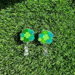 Peut inclure: Deux breloques trèfles à quatre feuilles vert et bleu avec des porte-badges rétractables argentés. Les breloques sont sur un fond de gazon vert.