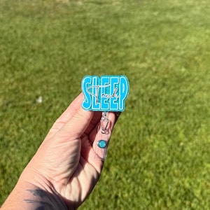 Può includere: Un porta badge turchese e bianco con le parole "Sleep Tech" in un carattere corsivo. Il porta badge ha una finitura glitterata e un cordino retrattile in plastica trasparente. Lo sfondo è un prato verde sfocato.