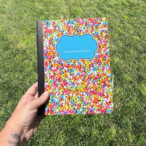 May include: A notebook with a black spine and a cover featuring a dense pattern of multicolored round sprinkles. Centered on the cover is a bright blue, decorative bracket-shaped label with a dotted line for writing a name or title.