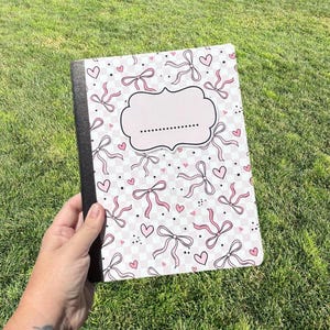 Puede incluir: Un cuaderno con lomo negro y una cubierta con un patrón de cuadros rosa claro y blanco, decorado con pequeños corazones rosas, puntos negros y lazos de cinta rosa. Una etiqueta central rosa claro con borde festoneado y línea punteada.