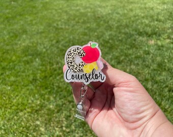 May include: A clear glitter badge holder with a colorful design featuring a leopard print letter C, a red apple, and a yellow and white striped design. The badge holder has a silver retractable badge reel and the text "Counselor" is printed on the front.