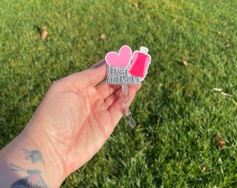 Puede incluir: Un carrete retráctil para placa con un corazón rosa y un diseño de maleta rosa. El texto "FLIGHT ATTENDANT" se muestra en letras plateadas. El carrete tiene un clip de plástico transparente y un clip de cinturón de metal plateado.
