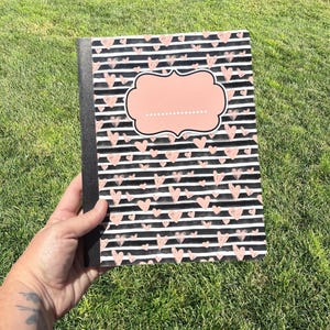 May include: A rectangular notebook featuring a pattern of horizontal black and white stripes interspersed with small pink hand-drawn hearts. A solid pink label with a decorative scalloped border and a dotted line is centered on the front cover.