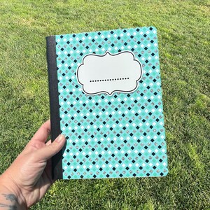 May include: A rectangular notebook featuring a teal, black, and white diamond pattern with silver glitter accents. The cover includes a central white label with a decorative black border and a dotted line for writing, finished with a black spine.