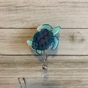 Puede incluir: Un carrete de identificación de tortuga marina azul con un cordón retráctil plateado. La tortuga es de color azul claro con un caparazón azul oscuro.