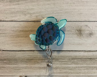 May include: A blue sea turtle badge reel with a silver retractable cord. The turtle is a light blue color with a dark blue shell.