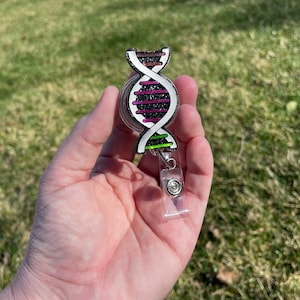 Könnte beinhalten: Ein schwarzer und weißer DNA-Doppelhelix-Badge-Rollenhalter mit rosa, grünen und braunen Akzenten. Der Rollenhalter ist aus Glitzer gefertigt und hat eine transparente, einziehbare Schnur aus Kunststoff.