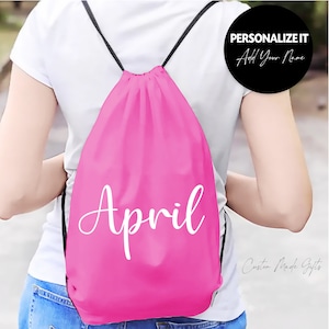 May include: Pink drawstring bag with the name "April" printed in white cursive font.  The bag is being worn by a person.  "Personalize It Add Your Name" is written in white text on a black circle.
