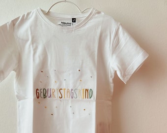 May include: White t-shirt with the text "Geburtstagskind" embroidered in rainbow colors.