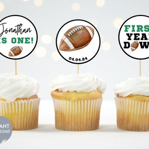 Puede incluir: Tres cupcakes con glaseado blanco cubiertos con toppers redondos de papel para cupcakes con un dise&ntilde;o de f&uacute;tbol. Los toppers tienen el texto "Jonathan is one!"  "First Year Down" y "04.04.24".