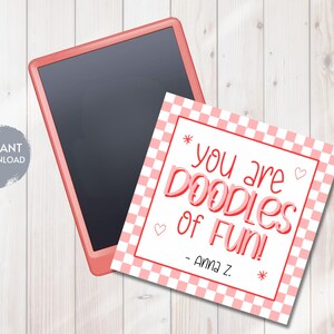 Könnte beinhalten: Ein digitales Tablet mit schwarzem Bildschirm und korallfarbenem Rahmen liegt neben einer quadratischen Karte. Die Karte hat ein rosa-weißes Karomuster und den Text "you are DOODLES of FUN!" mit einer Unterschrift.